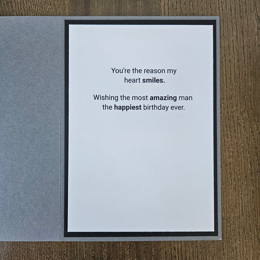 A square greeting card with a white center panel and black border, placed on a gray textured cover, photographed on a wood surface. The card reads: 'You're the reason my heart smiles.