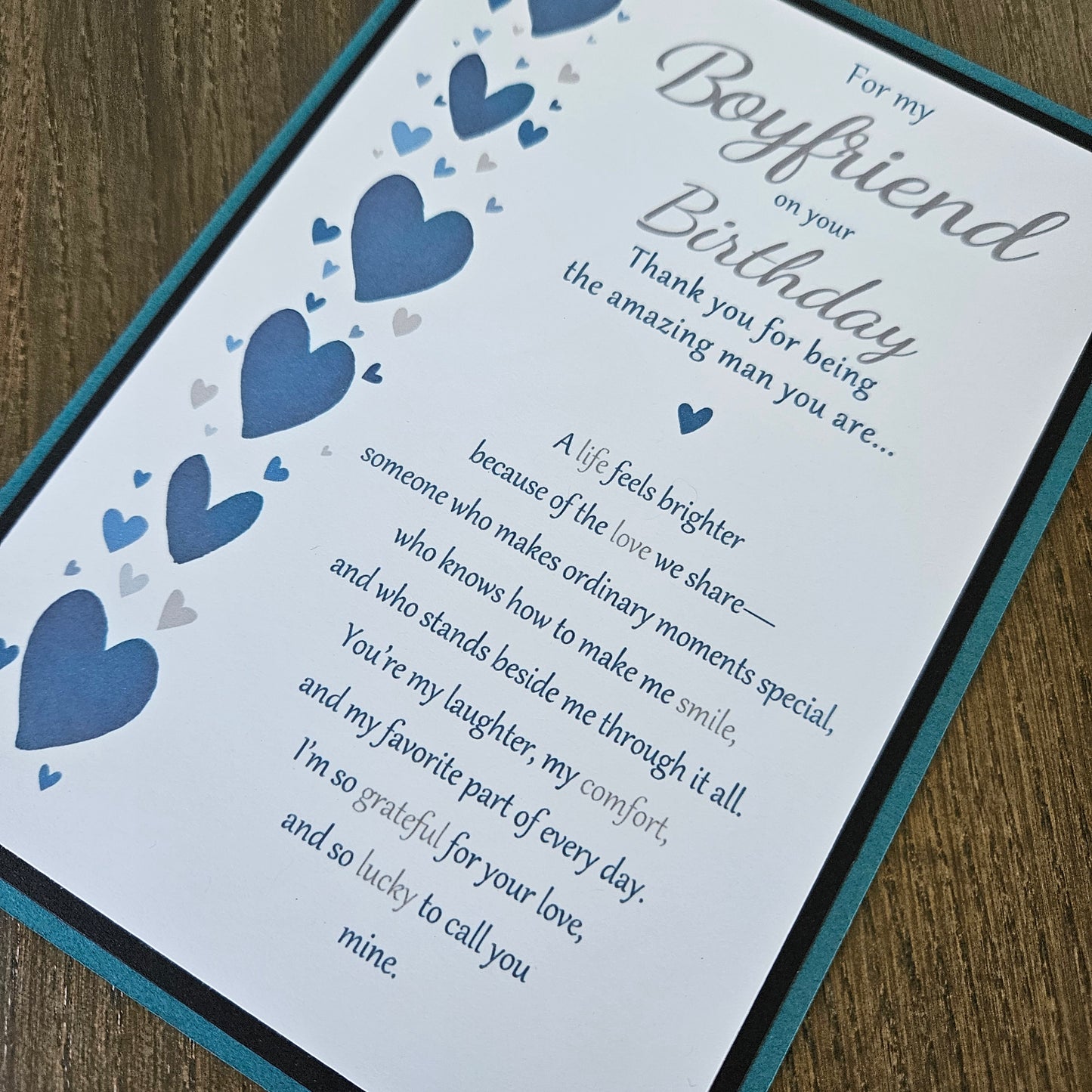 A close-up of a printed birthday card with a teal border, featuring blue and gray heart illustrations and a sentimental message addressed to a boyfriend, placed on a wooden surface.