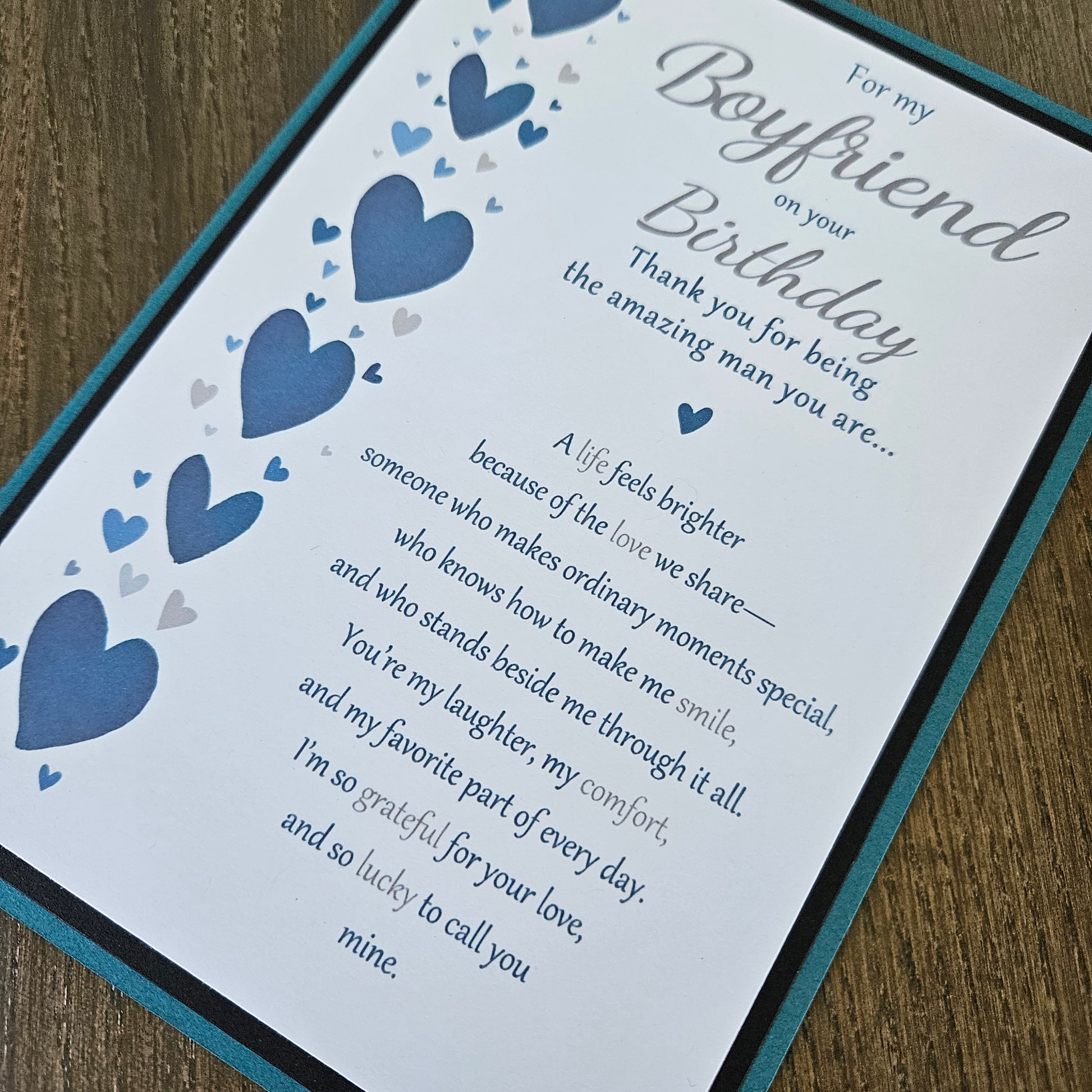 A close-up of a printed birthday card with a teal border, featuring blue and gray heart illustrations and a sentimental message addressed to a boyfriend, placed on a wooden surface.