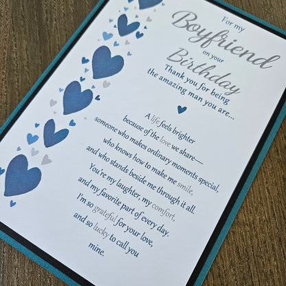 A close-up of a printed birthday card with a teal border, featuring blue and gray heart illustrations and a sentimental message addressed to a boyfriend, placed on a wooden surface.