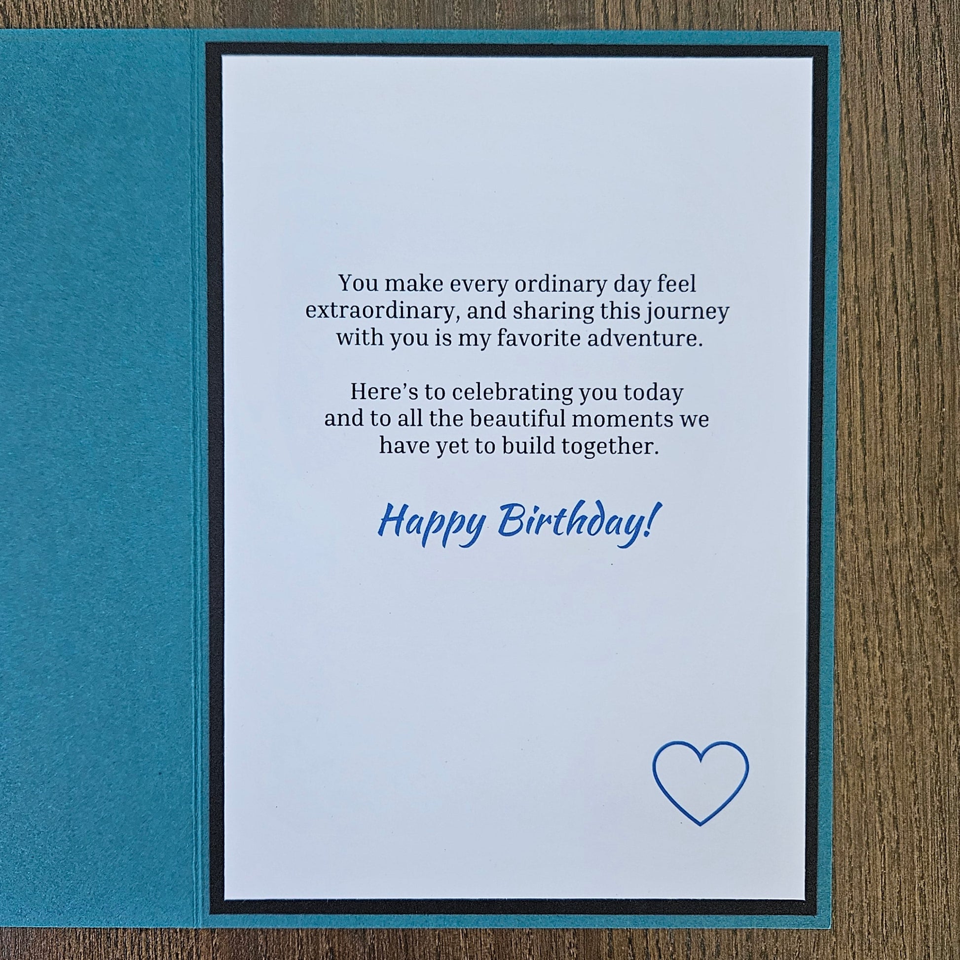 A square birthday card with a teal border and a white center panel, placed on a brown wood surface. The card has a printed message in black text with 'Happy Birthday!' in blue script and a small blue heart outline near the bottom right.