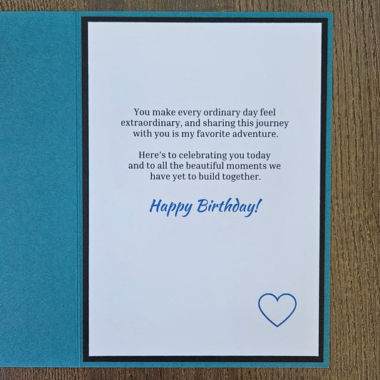 A square birthday card with a teal border and a white center panel, placed on a brown wood surface. The card has a printed message in black text with 'Happy Birthday!' in blue script and a small blue heart outline near the bottom right.