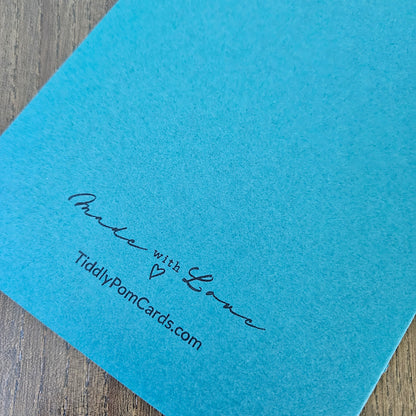 A close-up of a bright turquoise blue textured card or paper sheet placed on a wooden surface, with black printed text reading 'Pride with Love' and 'TiddlyPomCards.com'.