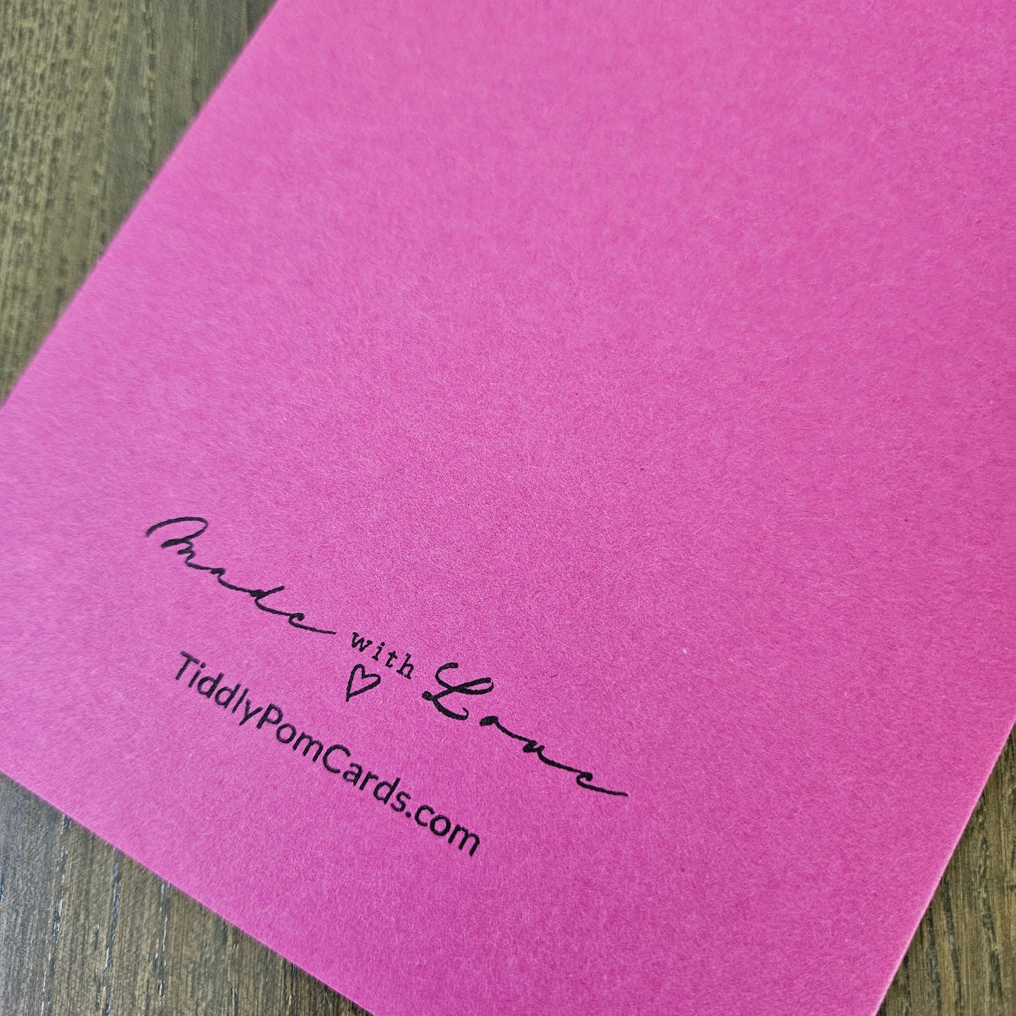 A bright pink pom pom card with black printed text reading 'Made with Love' and 'TiddlyPomCards.com' resting on a dark wood surface.