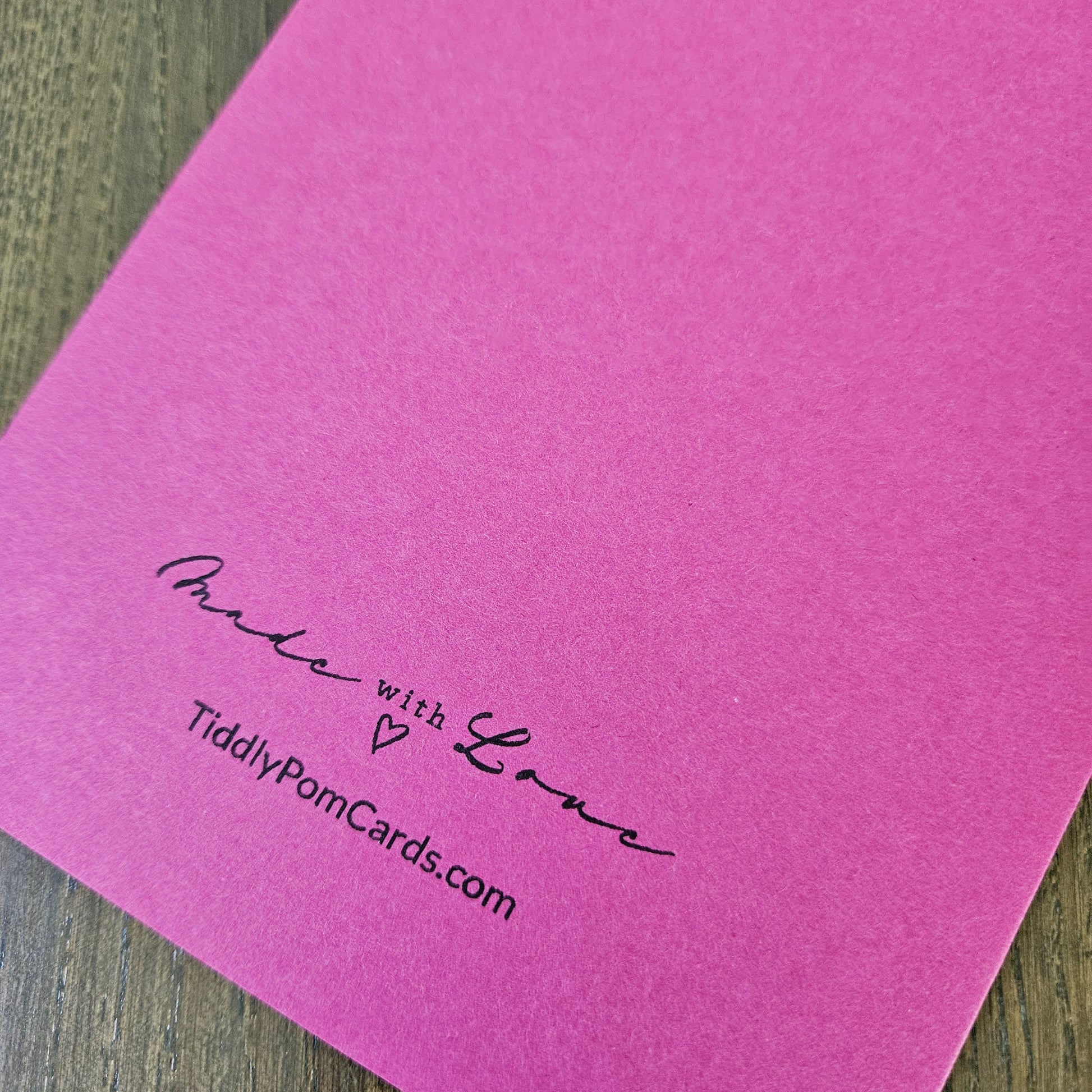 A bright pink pom pom card with black printed text reading 'Made with Love' and 'TiddlyPomCards.com' resting on a dark wood surface.