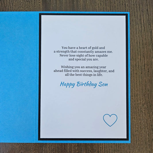 A birthday card for a son shown open on a dark wood surface, with a bright blue left panel and a white center panel containing a poem, the words 'Happy Birthday Son' in blue script, and a small blue heart outline.