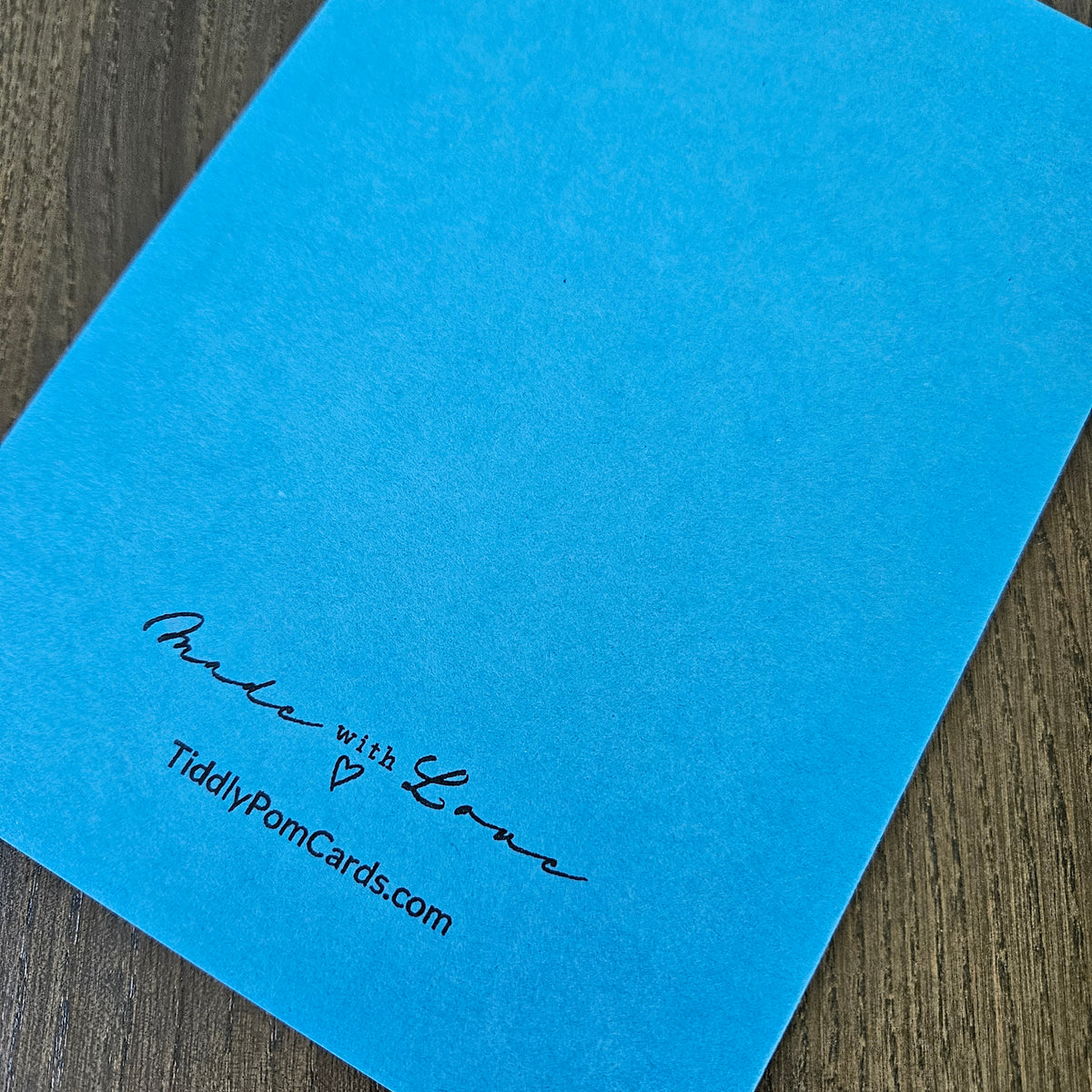 A bright blue textured card or envelope corner with black printed text reading 'Palm with Love' and 'TiddlyPomCards.com' on a dark wood surface.