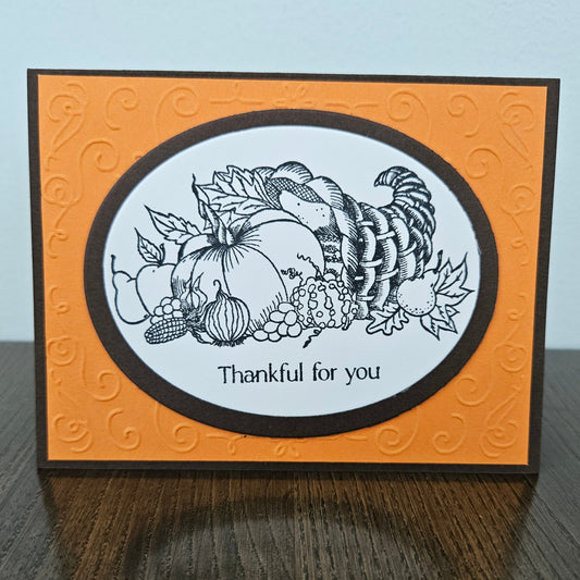 A handmade Thanksgiving greeting card standing on a dark wooden table against a light gray wall. The card features an orange embossed background with a central white oval framed by a black border, containing a black line art illustration of autumn harvest elements like a cornucopia, pumpkins, grapes, and a corn cob, with the text 'Thankful for you' below.