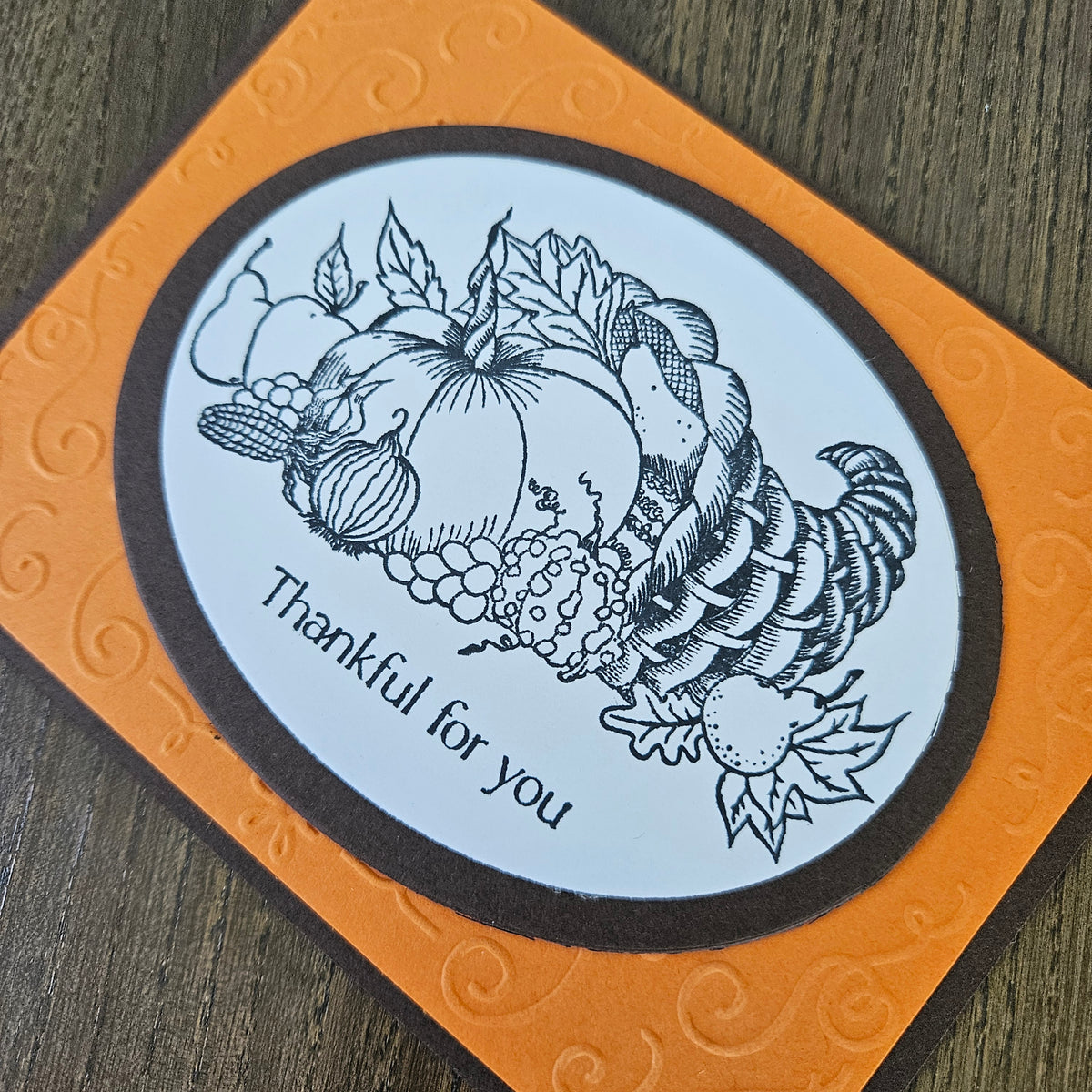 A square orange card with embossed swirls, featuring a central oval white panel with a black line-art illustration of a harvest cornucopia, pumpkins, grapes, and leaves, plus the words 'Thankful for you' on a wood surface.