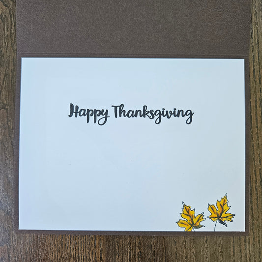 A square handmade Thanksgiving greeting card with a white front and brown backing, placed on a wooden surface. The front reads 'Happy Thanksgiving' in black script, with two small yellow maple leaves in the bottom right corner.