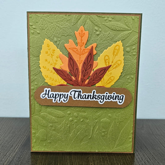 A handmade Thanksgiving greeting card featuring an embossed olive green background with layered autumn leaves in yellow, orange, and brown, and a brown banner reading 'Happy Thanksgiving' on a light blue background.