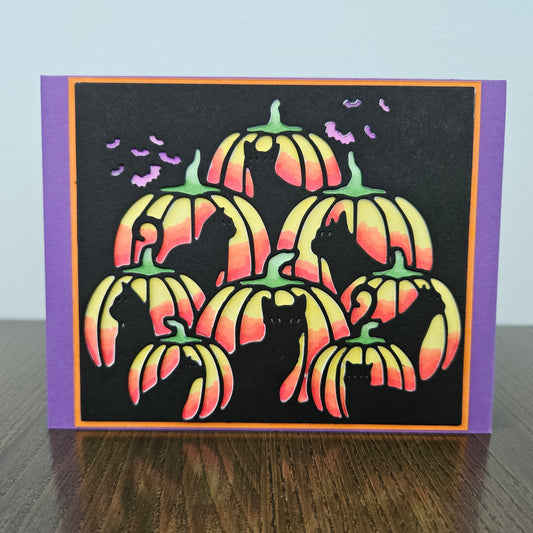 A handmade Halloween greeting card featuring silhouettes of cats inside striped pumpkins with orange, yellow, and green gradients on a black background, with purple bats and a purple border, displayed on a wooden surface.