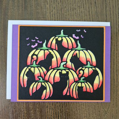 A square Halloween greeting card featuring stylized orange and pink pumpkins with black cat silhouettes, set against a black background with purple borders, photographed on a wooden surface.