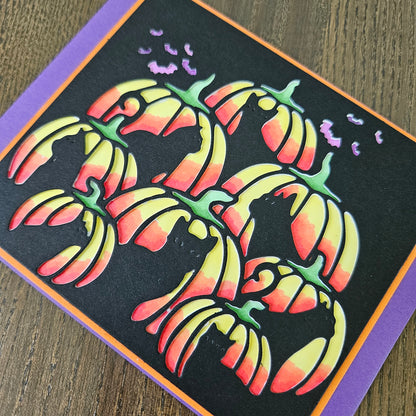 A close-up of a handmade Halloween card featuring stylized layered pumpkins in a yellow-to-orange gradient on a black background, with purple bats and a purple border.