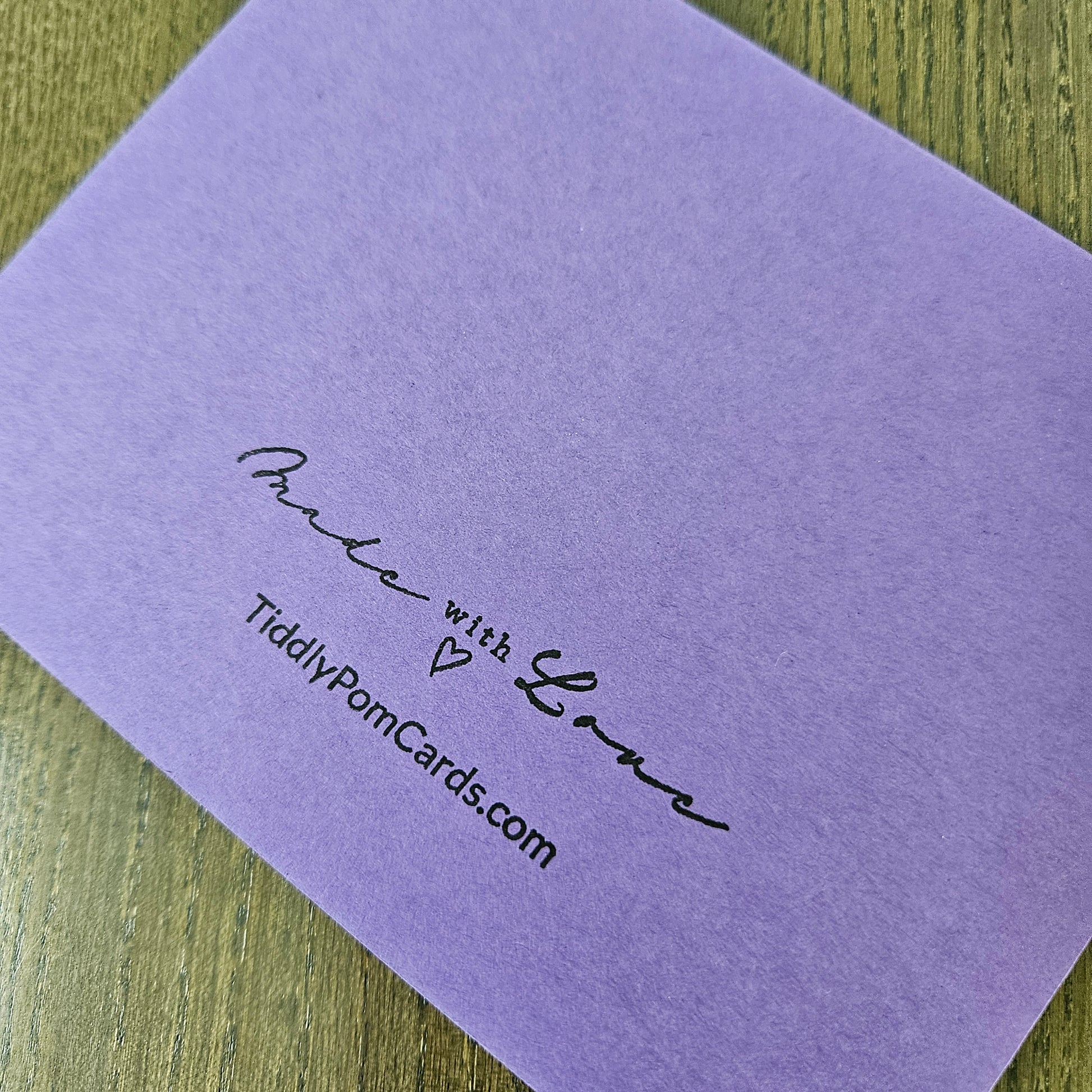 A lavender purple textured card with black printed text reading 'Made with Love' and 'TiddlyPomCards.com' resting on a wooden surface.