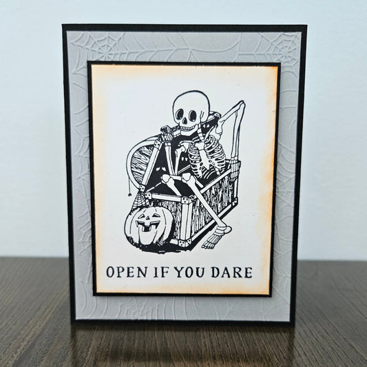 A framed Halloween greeting card or art print standing on a dark wooden surface against a light gray wall. The design shows a black-and-white skeleton sitting in a chair with a jack-o'-lantern and the text 'OPEN IF YOU DARE'.