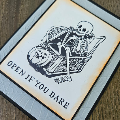 A square Halloween-themed art print or card featuring a black-and-white skeleton sitting in a chest with a jack-o'-lantern, with the text 'OPEN IF YOU DARE' on a warm off-white background, photographed on a wood surface.