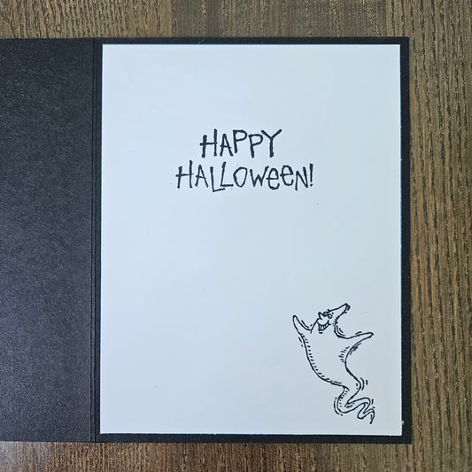 A handmade Halloween greeting card with a white front panel reading 'HAPPY HALLOWEEN!' in black lettering and featuring a small black line drawing of a ghost, displayed on a dark textured backing and a wooden surface.