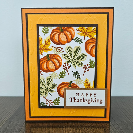 A handmade layered greeting card with autumn-themed stamping, featuring embossed orange and yellow borders, pumpkins, leaves, and a small banner that reads 'HAPPY Thanksgiving' on a wooden surface.