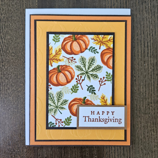 A handmade Thanksgiving greeting card featuring a layered autumn design with orange pumpkins, green leaves, and yellow maple leaves on a white background, framed by orange and yellow borders with a small 'HAPPY Thanksgiving' banner, photographed on a dark wood surface.
