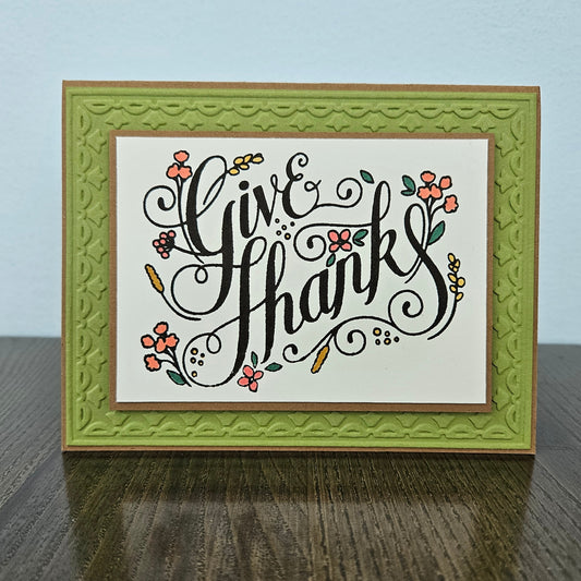 A handmade greeting card with the words 'Give Thanks' in black cursive script, decorated with small orange and yellow flowers and green leaves. The card is layered with a textured olive green border and a brown outer frame, standing on a dark wooden table against a light blue wall.
