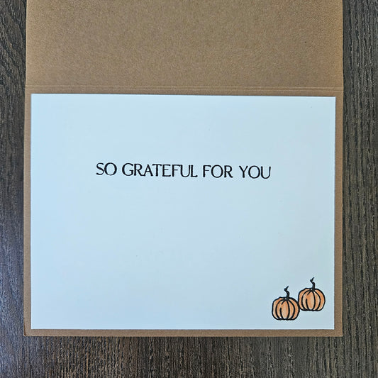 A square greeting card with a kraft envelope, featuring the printed text 'SO GRATEFUL FOR YOU' and two small orange pumpkins in the bottom right corner, displayed on a dark wood background.