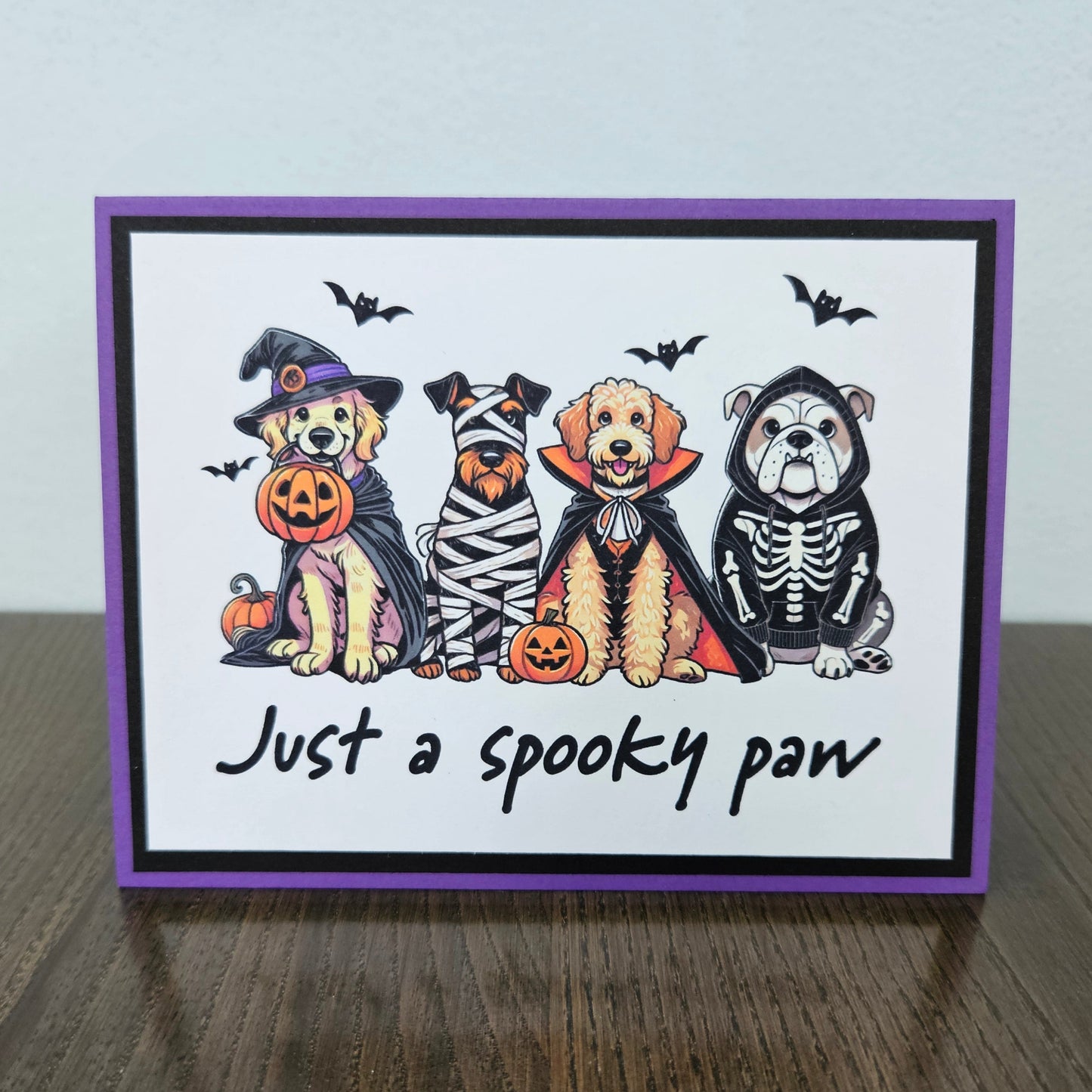 Front of a handmade A2 Halloween card featuring four dogs in Halloween costumes framed with black and purple borders with text reading Just a spooky paw.