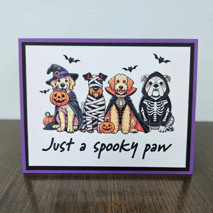Front of a handmade A2 Halloween card featuring four dogs in Halloween costumes framed with black and purple borders with text reading Just a spooky paw.