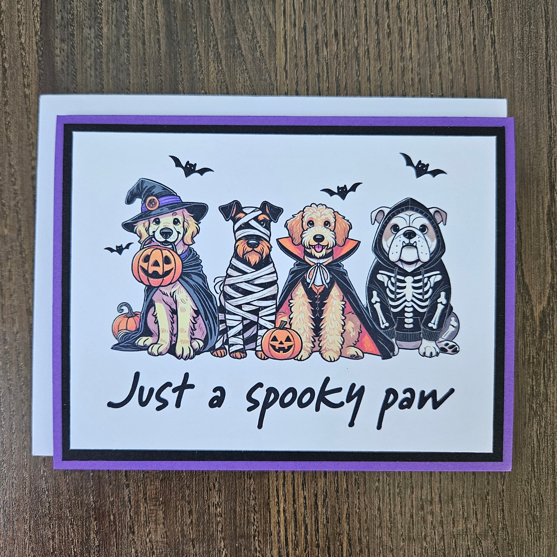 Front of a handmade A2 Halloween card featuring four dogs in Halloween costumes framed with black and purple borders with text reading Just a spooky paw.