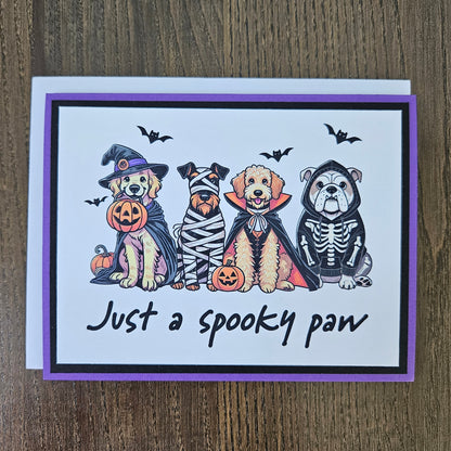 Front of a handmade A2 Halloween card featuring four dogs in Halloween costumes framed with black and purple borders with text reading Just a spooky paw.