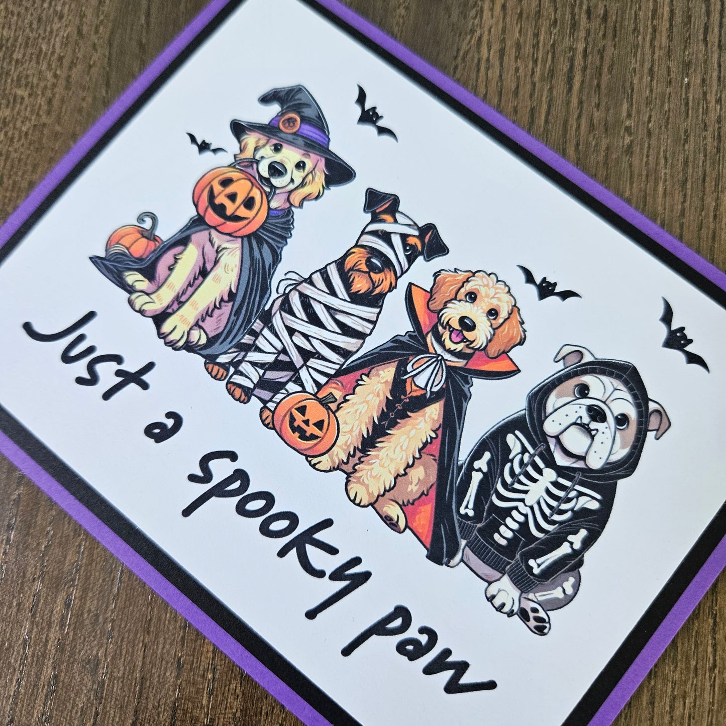 Front of a handmade A2 Halloween card featuring four dogs in Halloween costumes framed with black and purple borders with text reading Just a spooky paw.