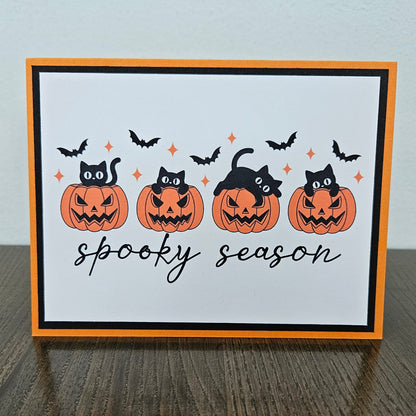 Front of a handmade A2 Halloween card featuring four black cats with jack-o'-lantern pumpkins and flying bats framed in black and orange borders with text reading spooky season.