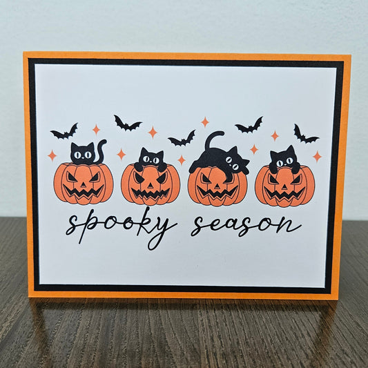 Front of a handmade A2 Halloween card featuring four black cats with jack-o'-lantern pumpkins and flying bats framed in black and orange borders with text reading spooky season.