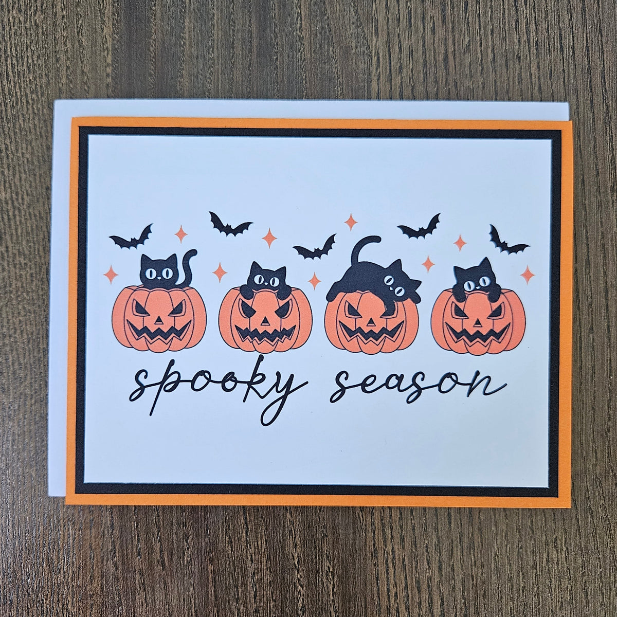 Front of a handmade A2 Halloween card featuring four black cats with jack-o'-lantern pumpkins and flying bats framed in black and orange borders with text reading spooky season.