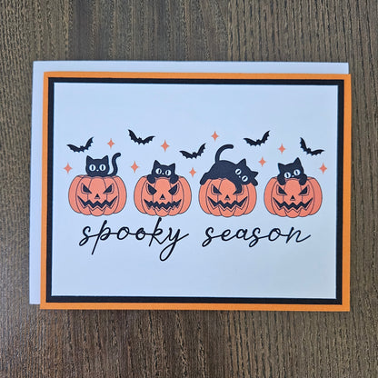 Front of a handmade A2 Halloween card featuring four black cats with jack-o'-lantern pumpkins and flying bats framed in black and orange borders with text reading spooky season.