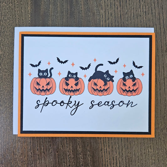 Front of a handmade A2 Halloween card featuring four black cats with jack-o'-lantern pumpkins and flying bats framed in black and orange borders with text reading spooky season.