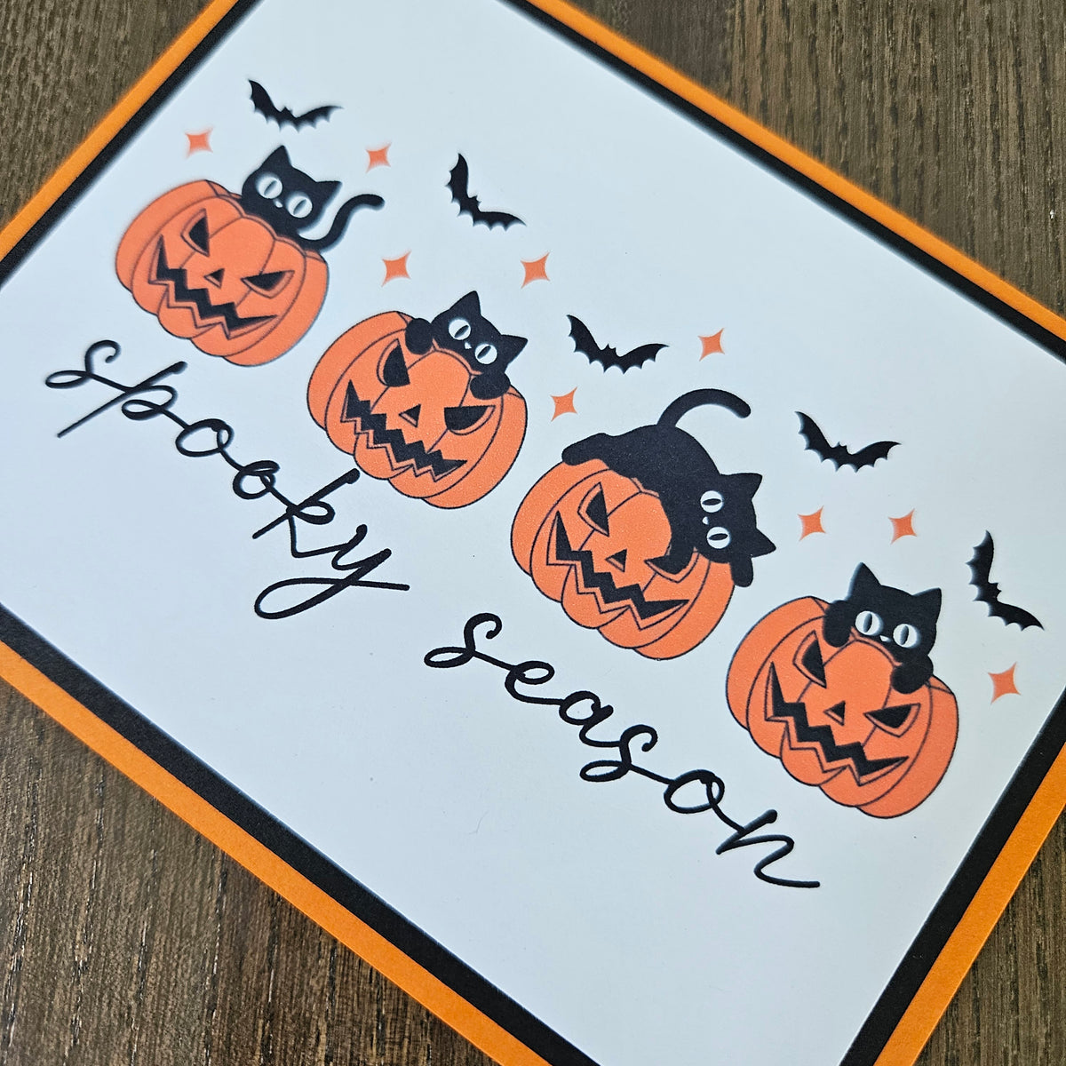A square Halloween greeting card with a white background, featuring four orange jack-o'-lantern pumpkins with black cats, black bats, and the words 'spooky season' in black script. The card has an orange and black border and is photographed on a wooden surface.
