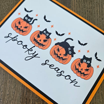 A square Halloween greeting card with a white background, featuring four orange jack-o'-lantern pumpkins with black cats, black bats, and the words 'spooky season' in black script. The card has an orange and black border and is photographed on a wooden surface.