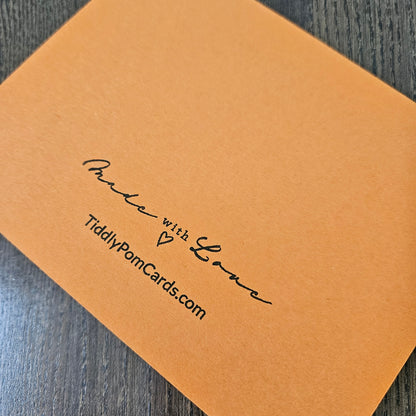 A close-up of an orange pom pom card with black printed text reading 'Made with Love' and 'TiddlyPomCards.com' on a dark wood surface.