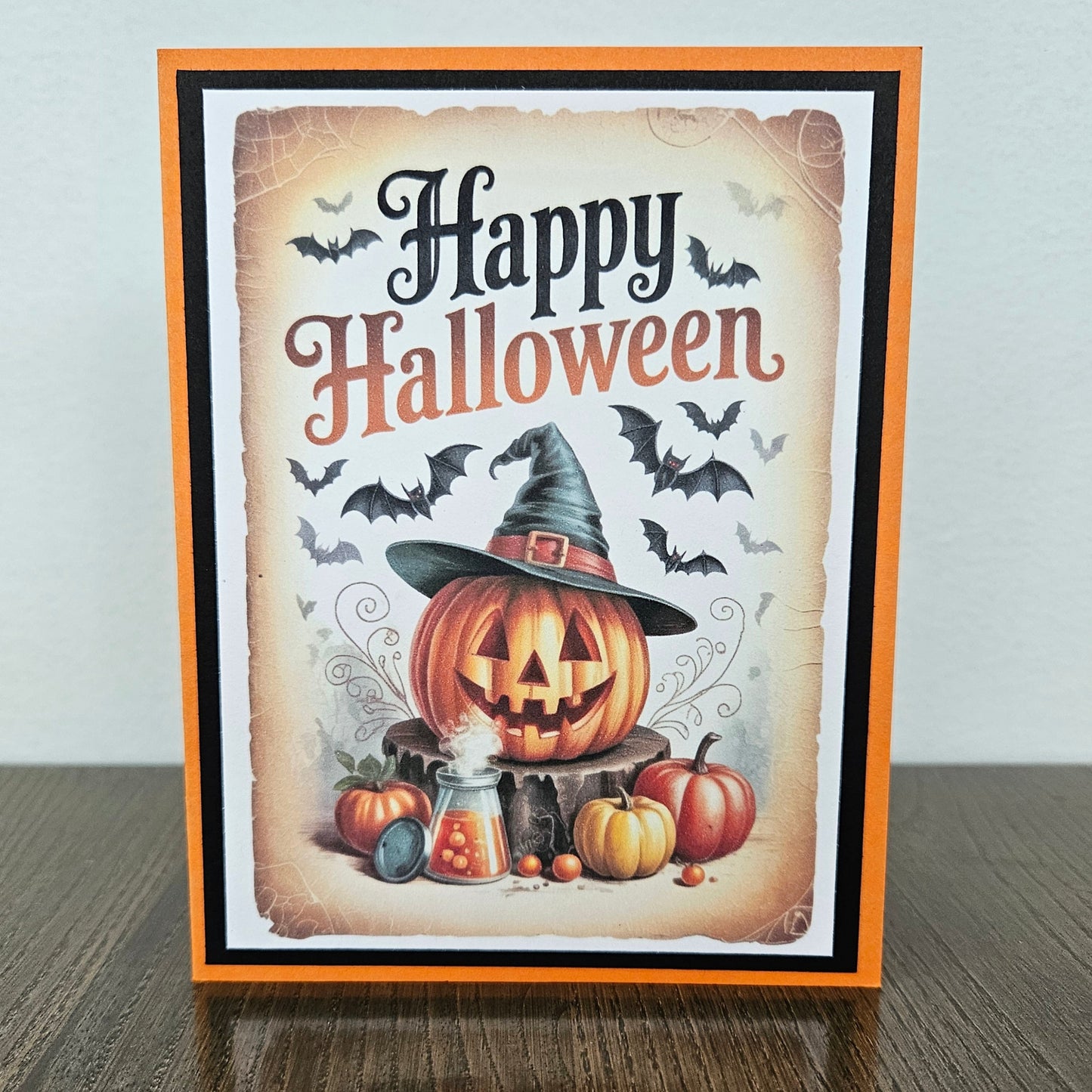 Front of a handmade A2 Halloween card featuring a jack-o'-lantern wearing a witch hat with flying bats and pumpkins framed by black and orange borders with text reading Happy Halloween.