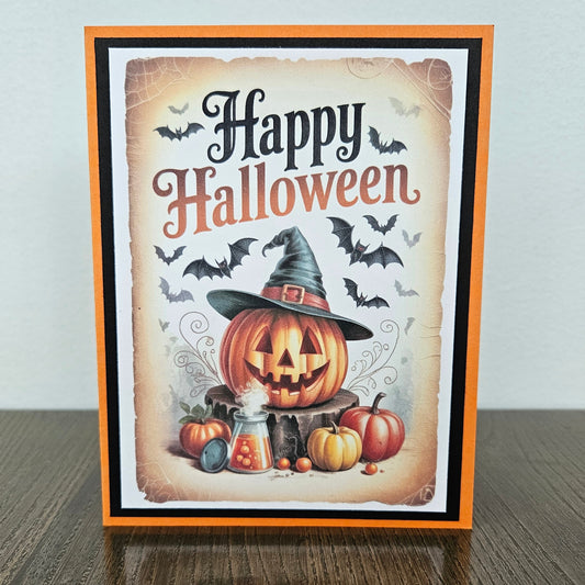 Front of a handmade A2 Halloween card featuring a jack-o'-lantern wearing a witch hat with flying bats and pumpkins framed by black and orange borders with text reading Happy Halloween.