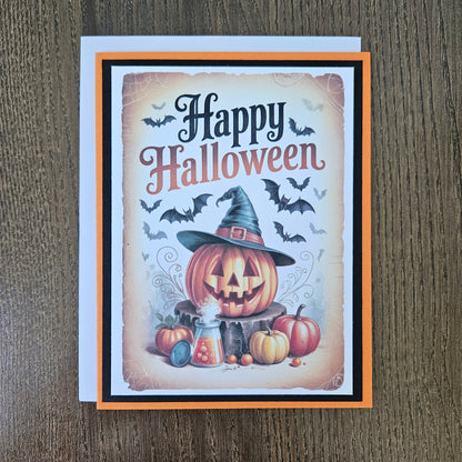 A square Halloween greeting card with a white envelope, placed on a dark wood surface. The card features a vintage-style illustration of a carved pumpkin wearing a witch hat, surrounded by smaller pumpkins, bats, and the words 'Happy Halloween' in black and peach lettering.