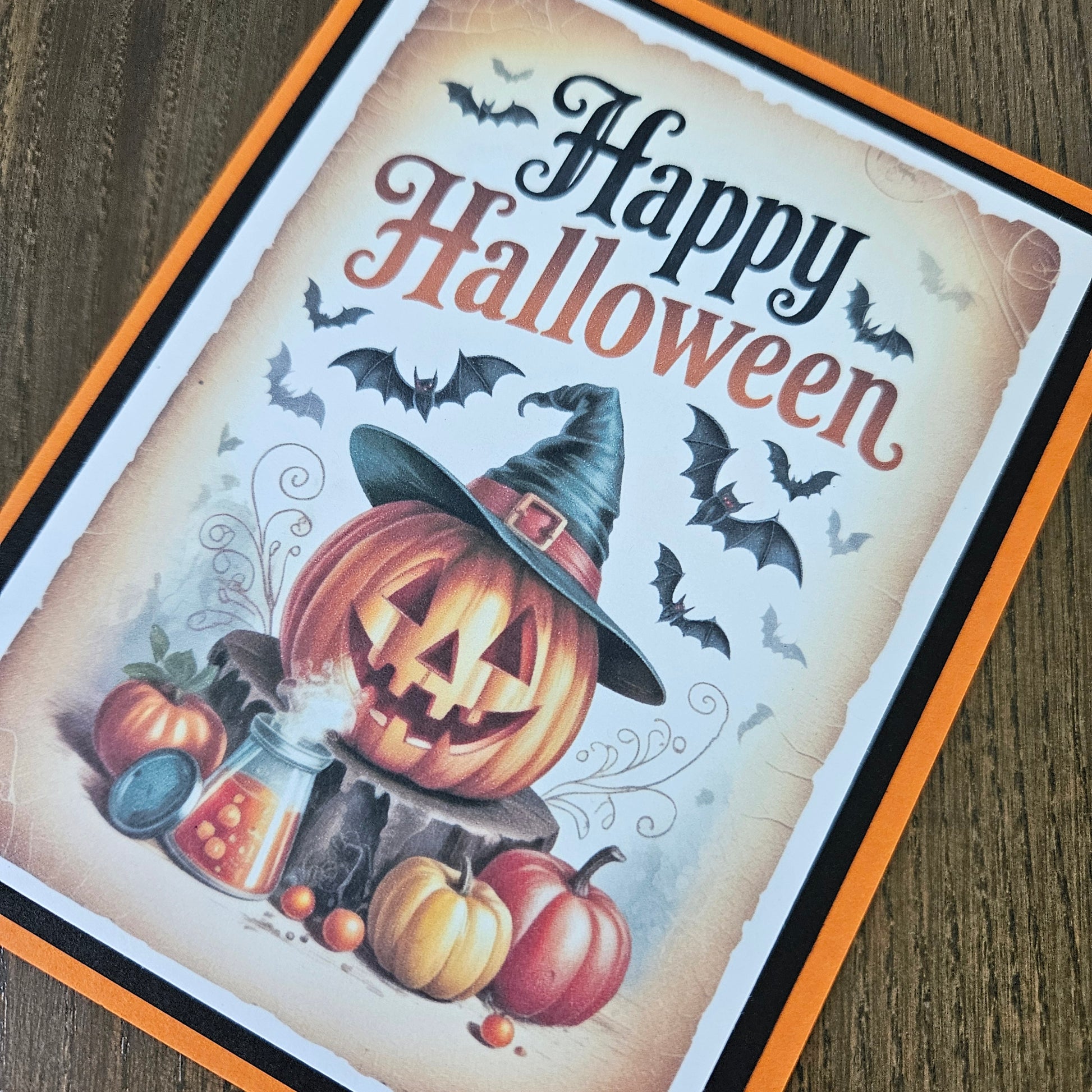 A square Halloween greeting card featuring a carved pumpkin, witch hat, pumpkins, bats, and the words “Happy Halloween” in decorative lettering, photographed on a wood surface.