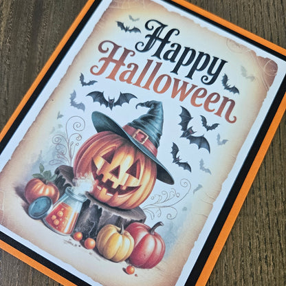A square Halloween greeting card featuring a carved pumpkin, witch hat, pumpkins, bats, and the words “Happy Halloween” in decorative lettering, photographed on a wood surface.
