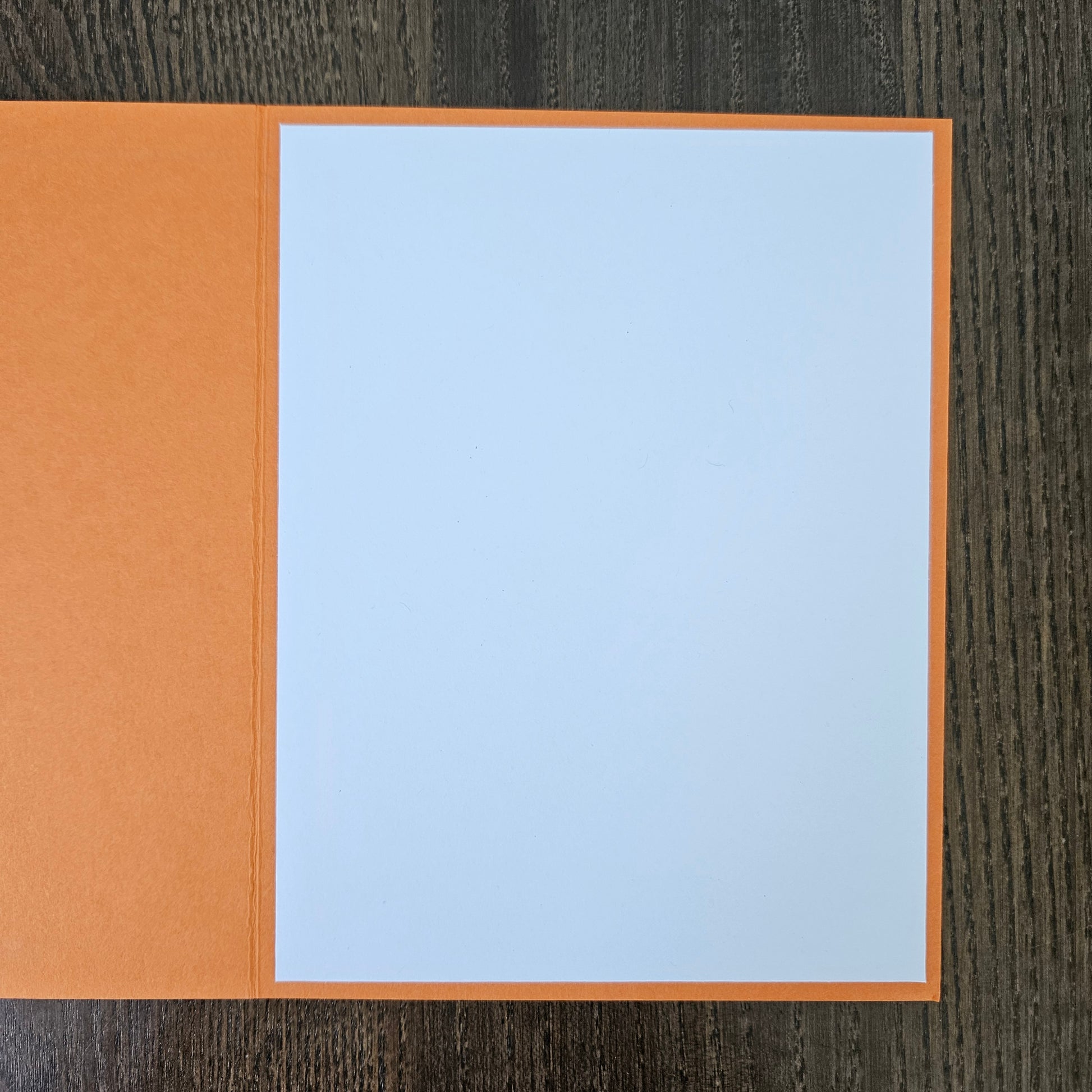An open orange greeting card with a blank light blue insert page, displayed on a dark wood background.