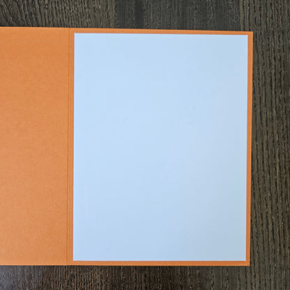 An open orange greeting card with a blank light blue insert page, displayed on a dark wood background.