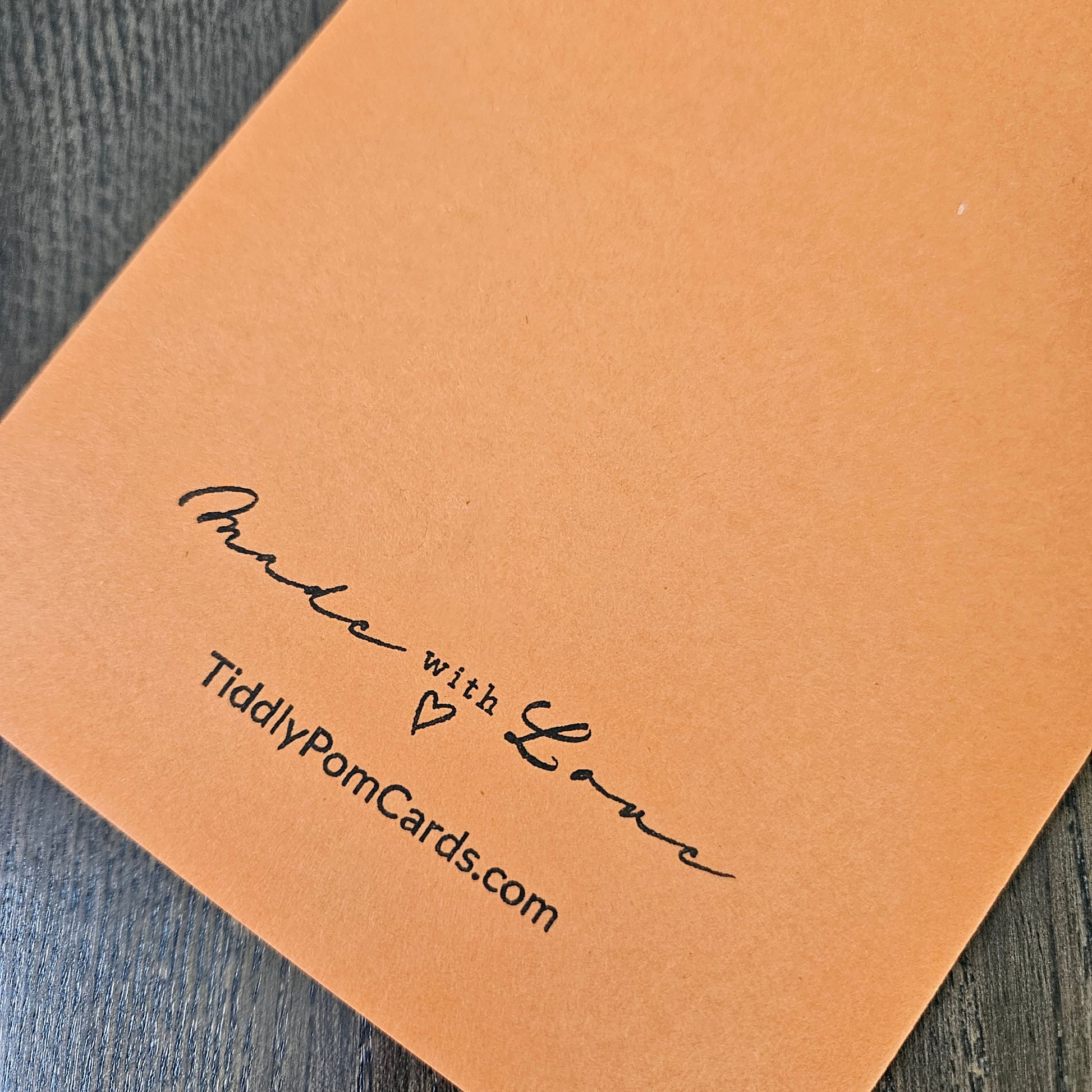 A close-up of a square kraft paper card with black printed text reading 'Made with Love' and 'TiddlyPomCards.com' on a dark wood surface.