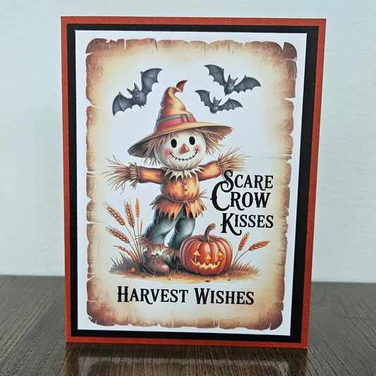 A handmade Halloween greeting card featuring a smiling scarecrow, a carved pumpkin, bats, and the text 'SCARE CROW KISSES' and 'Harvest Wishes', displayed standing on a dark wooden surface against a light gray wall.