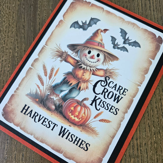 A square Halloween greeting card featuring a smiling scarecrow, a carved pumpkin, bats, wheat stalks, and the text 'Scare Crow Kisses' and 'Harvest Wishes,' photographed on a wooden surface.