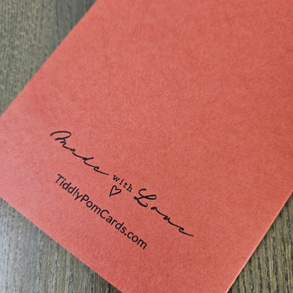 A close-up of a coral pink textured card or envelope corner with black printed text reading 'Anita with Love' and 'TiddlyPomCards.com' on a wood surface.
